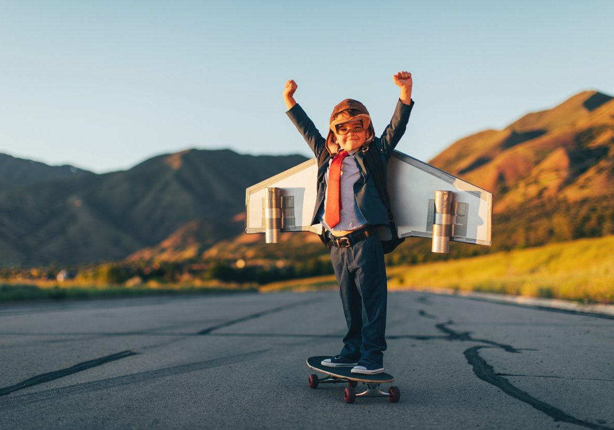 Young boy businessman wearing flight goggles and homemade jet pack stands with arms raised in accomplishment on a skateboard ready to take off flying and launch his new business into the land of profits and growth. He is dressed in a business suit. Image taken in Utah, USA.